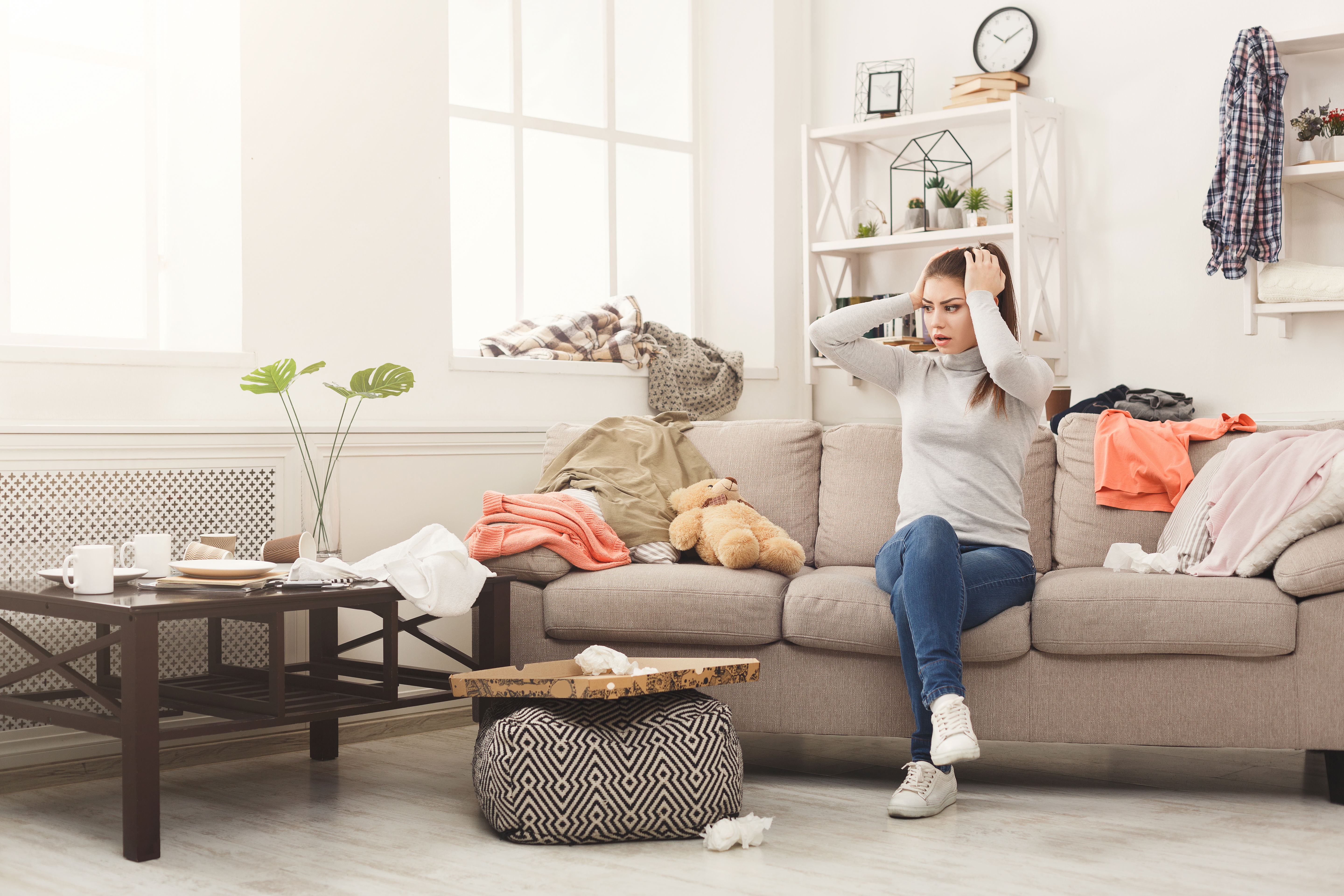 Desperate helpless woman sitting on sofa in messy living room. Young girl surrounded by many stack of clothes. Disorder and mess at home.
