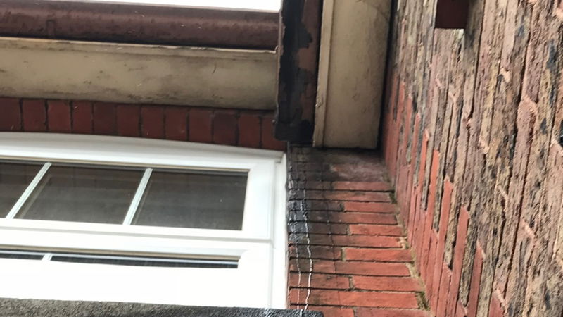 Water leaking from a gutter onto a masonry wall, mould visible.